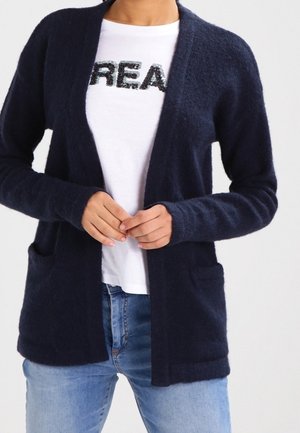 Cárdigan azul marino hecho de una suave tela texturizada, con frente abierto, mangas largas y dos bolsillos frontales, usado sobre una camisa blanca.