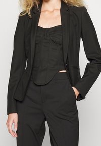 Blazer noir ajusté porté sur un corset noir cintré, avec un design structuré, une texture lisse et une légère brillance.