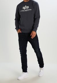 Felpa grigia scura con logo "ALPHA INDUSTRIES", abbinata a jeans scuri con accenni di spruzzi e sneakers bianche. Outfit casual.