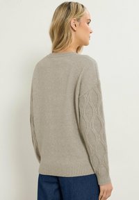 Lys grå sweater med rund halsudskæring, ribbede manchetter og et struktureret kabelstrik mønster på ærmerne. Blødt, strikket stof.