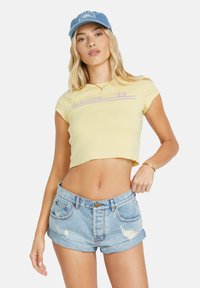 T-shirt cropped jaune avec des rayures de logo violettes associé à un short en denim bleu clair déchiré. Casquette bleue avec accent de logo.