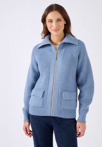 Cárdigan de punto azul claro con textura de canalé, cuello oversized, cremallera dorada y dos bolsillos frontales.