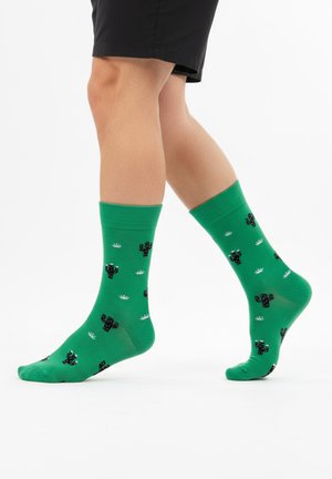 Calcetines verdes con patrones de cactus en negro, con detalles blancos y puño acanalado. La tela parece suave y elástica.