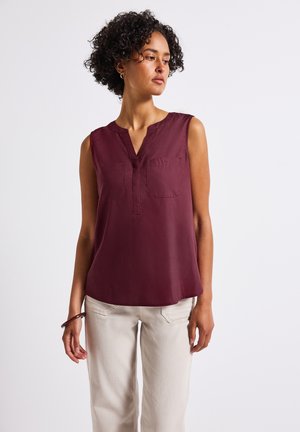Top bordeaux senza maniche con scollo a V, patta frontale e due tasche sul petto; tessuto leggero con una texture morbida; abbinato a pantaloni beige.