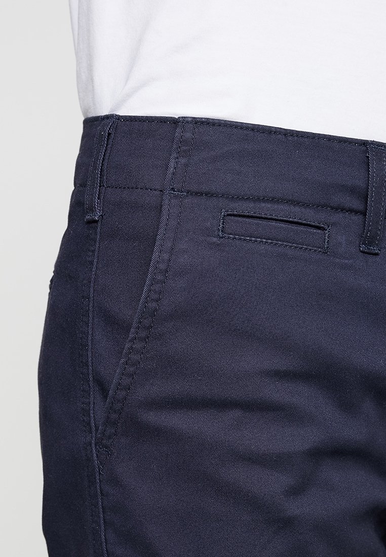 Gros plan sur la ceinture et la zone de la poche d'un pantalon bleu marine porté avec une chemise blanche rentrée, montrant les détails de la couture et les passants de ceinture.