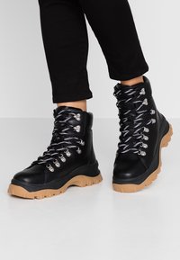 Botas de senderismo de cuero negro con ojales plateados, cordones en negro y blanco, y una suela de goma beige gruesa. Adecuadas para actividades al aire libre.