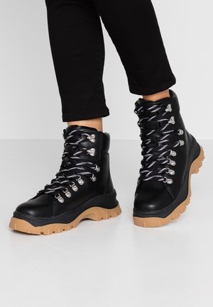 Botas de senderismo de cuero negro con ojales plateados, cordones en negro y blanco, y una suela de goma beige gruesa. Adecuadas para actividades al aire libre.