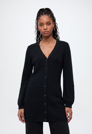 Femme aux cheveux tressés portant un long cardigan noir boutonné avec un col en V et de longues manches, debout devant un fond clair uni.