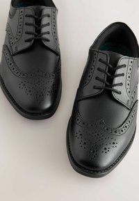 Chaussures habillées en cuir noir avec un design brogue, ornées de perforations décoratives, à lacets plats et une pointe arrondie. Présence de contrastes de texture subtils.