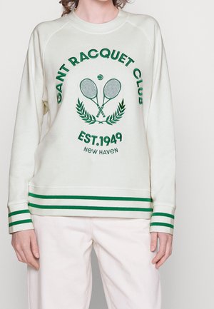 Osoba nosząca kremową bluzę z zielonym logo rakiety tenisowej oraz napisem „GANT Racquet Club EST. 1949 New Haven”, zestawioną z kremowymi spodniami.