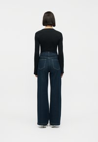 Γυναίκα με εφαρμοστή μαύρη μακρυμάνικη μπλούζα, ψηλόμεσο φαρδύ παντελόνι τζιν σκούρου μπλε και λευκά αθλητικά παπούτσια, στέκεται με την πλάτη στο βλέμμα.