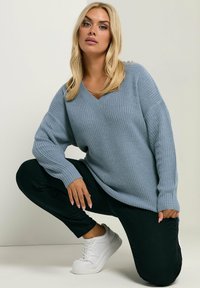 Jasnoniebieski sweter dzianinowy z dekoltem w kształcie litery V i prążkowaną strukturą, zestawiony z dopasowanymi czarnymi spodniami i białymi trampkami. Modelka pozuje w pozycji kucznej.