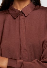 Gros plan sur une personne portant une chemise brune à col avec des manches longues, boutonnée.