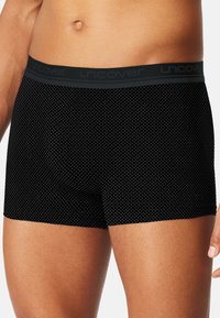 Schwarze Boxershorts mit einem geometrischen Muster, aus weichem Stoff, mit einem grauen elastischen Bund, auf dem das Wort "uncover" in wiederholendem Design zu sehen ist.