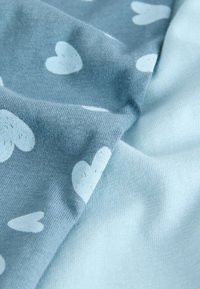 Tissu bleu clair présentant une texture douce avec des cœurs blancs éparpillés et des détails subtils. Le matériau semble extensible et confortable.