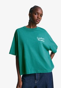 Grøn oversized bomulds t-shirt med korte ærmer, der har hvid tekst "Sted: Ferie" på venstre brystområde, parret med mørk denim jeans.