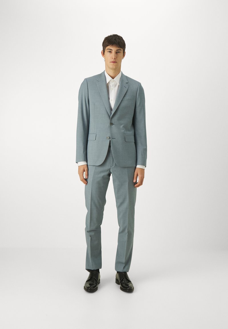 Costum bleu deschis, croit cu un fit slim, cu reveri tăiate, două buzunare frontale și pantaloni asortați; combinat cu o cămașă albă și pantofi negri.