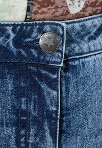 Mørkeblå denim stof med et falmet mønster. Har en sølvmetal knap med et stjernedesign og kontraststing langs taljen.