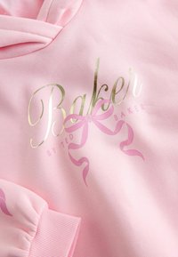 Rosa Kapuzenpullover mit einer weichen Textur, ausgestattet mit auffälligem goldenen "Baker"-Text und einer pinken Schleifen-Grafik. Rippbündchen betonen das Design.