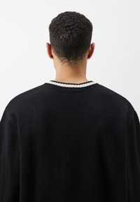 Czarny sweter z okrągłym dekoltem i białymi ściągaczami u kołnierza. Dzianinowa faktura, luźny krój i widoczne szwy na ramionach.