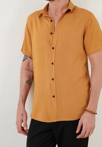 Koszula na krótki rękaw w kolorze musztardowym, wykonana z gładkiej tkaniny, z czarnymi guzikami i luźnym krojem.