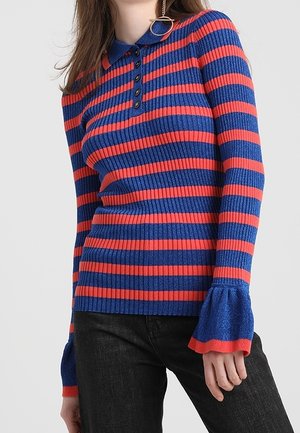 Vrouw draagt een blauw-rood horizontaal gestreepte geribbelde trui met kraag, knoopsluiting en uitlopende manchetten, gecombineerd met een zwarte broek.
