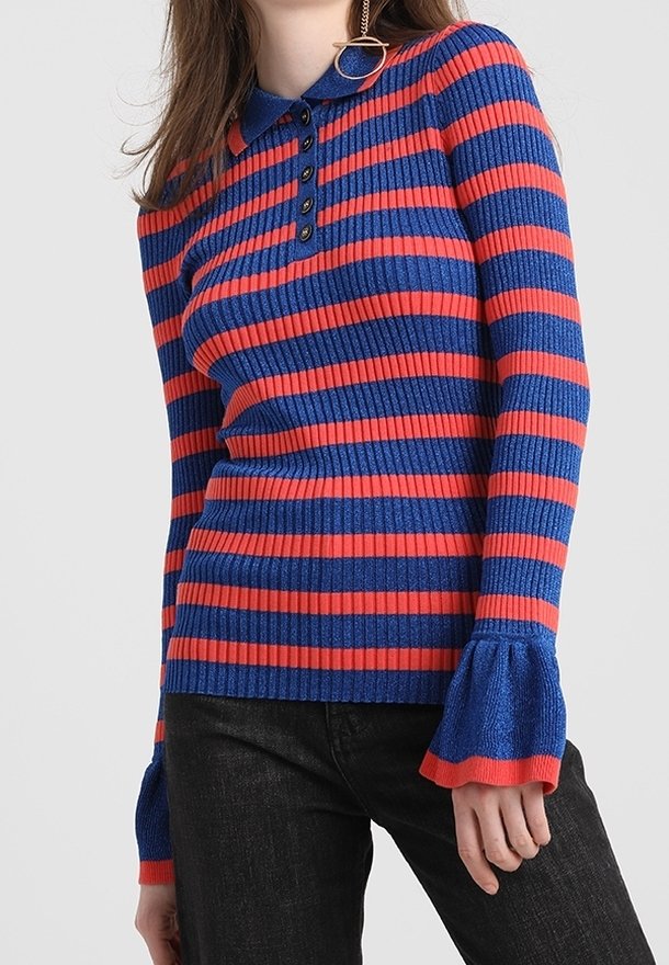 Femme portant un pull côtelé à rayures horizontales bleu et rouge avec un col, une patte de boutonnage et des poignets évasés, assorti à un pantalon noir.