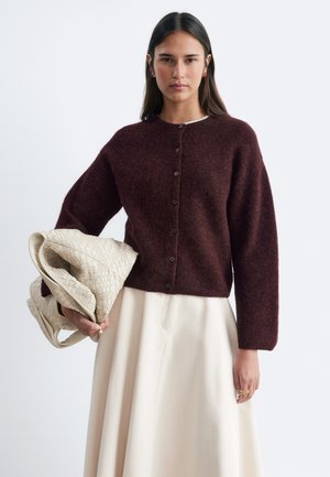 Donna con lunghi capelli scuri indossa un maglione marrone con bottoni e una gonna crema, tenendo una grande borsa intrecciata color crema su uno sfondo bianco semplice.