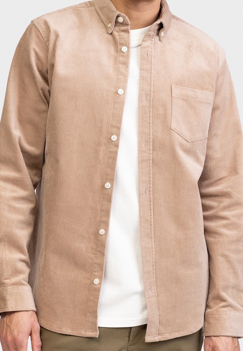 Man in een beige corduroy knoopshirt over een wit t-shirt en olijfgroene broek, staand tegen een effen achtergrond.