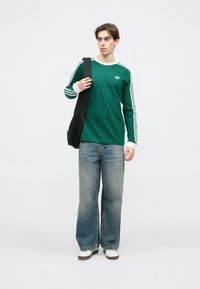 Zelená košeľa s dlhým rukávom s bielymi pruhmi, s malým logom; v kombinácii s svetlomodrými voľnými džínsami a bielymi teniskami. Čierna taška cez rameno.