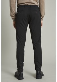 Svarta skräddarsydda byxor med slim fit, med baktill fickor med knappar, matchade med svarta skor. Tyget verkar slätt och hållbart.