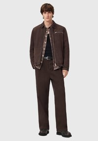 Brązowa kurtka z korduroyu z kieszenią na zamek, nałożona na koszulę w kratę, połączona z pasującymi brązowymi spodniami i czarnymi butami.