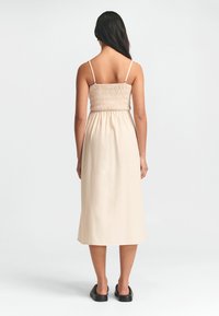 Maxi-kjole i lys beige med smock-buste, tynde stropper og en let skørt. Glat tekstur, midt på læggen længde. Sorte slip-on sko.