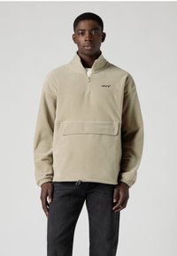 Smėlinės spalvos flisinis megztinis su pusiau užtraukiamu užtrauktuku, priekinė kišenė ir elastiniais rankogaliais. Ant krūtinės yra subtilus logotipas, o apačioje – reguliuojama raišteliu.