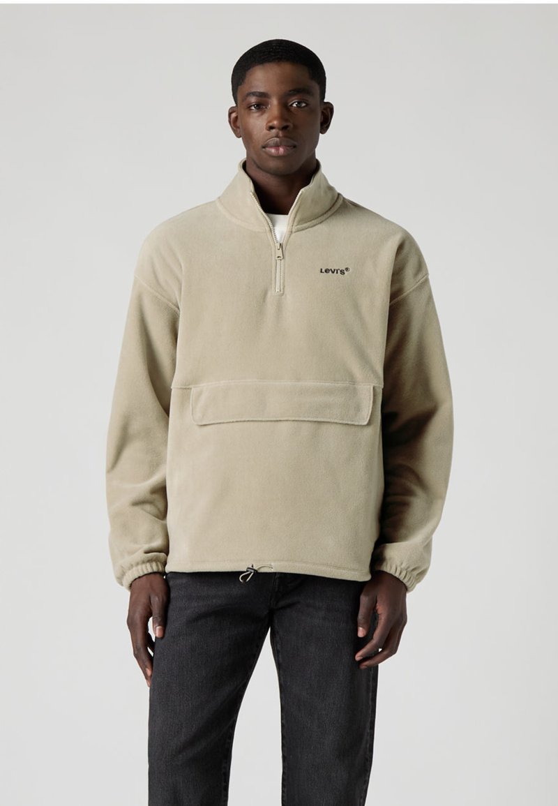 Beige fleecetröja med halvlås, framficka och elastiska muddar. Har en diskret logga på bröstet och justerbar dragsko i nederkanten.