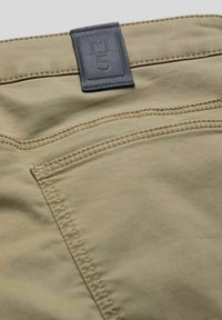 Beige Baumwollhose mit einem Lederlabel M5, sichtbaren Nähten und einem Detail mit einer Gesäßtasche. Glatte Textur mit einem lässigen Schnitt.