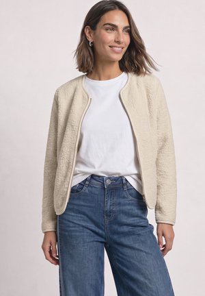 Femme portant une veste beige texturée zippée sur une chemise blanche et un jean bleu, souriante et regardant légèrement sur le côté.