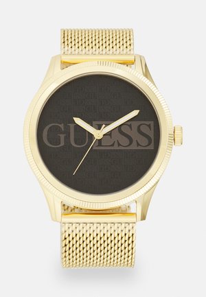 Montre dorée avec un boîtier rond et un bracelet en maille. Le cadran noir présente le logo "GUESS" en relief. Aiguilles dorées et détail de couronne.