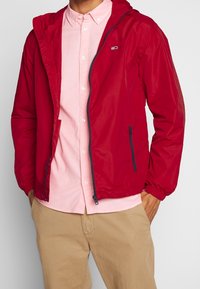 Man med en röd zip-up jacka över en ljusrosa skjorta med knappar och beigefärgade byxor, med händerna i jackfickorna, överkroppen synlig.