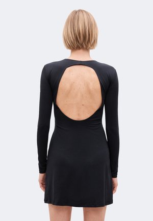 Femme aux cheveux blonds courts portant une robe noire à manches longues avec une grande découpe dans le dos, debout devant un fond clair uni.