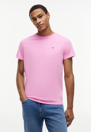 Ung mand iført en ensfarvet lyserød kortærmet T-shirt og blå jeans, stående med den ene hånd i lommen mod hvid baggrund.