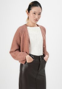 Mlada žena s rukama u džepovima nosi ružičasti pleteni kardigan, bijelu teksturiranu bluzu i crnu kožnu suknju visokog struka.