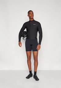 Svart sykkeltrøye med lange ermer og glidelås, kombinert med svarte shorts. Modellen holder en svart hjelm og har på seg svarte sykkelsko.