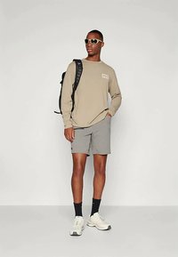 Beżowa koszulka z długim rękawem z logo, szare szorty, czarne skarpetki, białe sneakersy i okulary przeciwsłoneczne; nosi czarny plecak na lewym ramieniu.