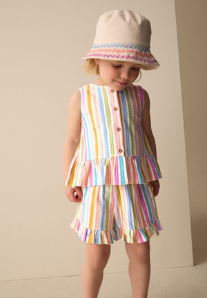 Junges Kind trägt ein ärmelloses Oberteil mit pastellfarbenen Regenbogenstreifen und Knöpfen sowie passende Rüschen-Shorts und einen beigen Hut mit buntem Rand.