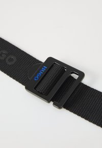 Cintura in tessuto nera con fibbia rettangolare in metallo nero. Presenta il marchio blu "HUGO" sulla fibbia. Superficie testurizzata, lunghezza regolabile.