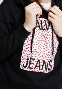 Sudadera negra con un frente de patrón floral en blanco que presenta el texto "CALVIN JEANS" en letras negras en negrita. Se ven los cordones.