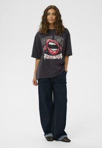 Göndör hajú nő fekete, nagy méretű grafikás pólóban, sötét bő szárú farmernadrágban és fekete fonott cipőben áll egy fehér háttér előtt.