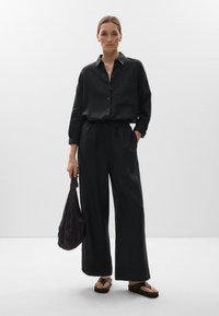 Fekete gombos ing zsebbel, laza fekete nadrág elasztikus derékkal és egy fekete, laza táska. Lapostalpú szandálok teszik teljessé a megjelenést.