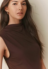 Bruine mouwloze top met een hoge halslijn. Zachte, gladde textuur met gedrapeerde stofdetails. Model heeft lang haar en gouden hoepel oorbellen.